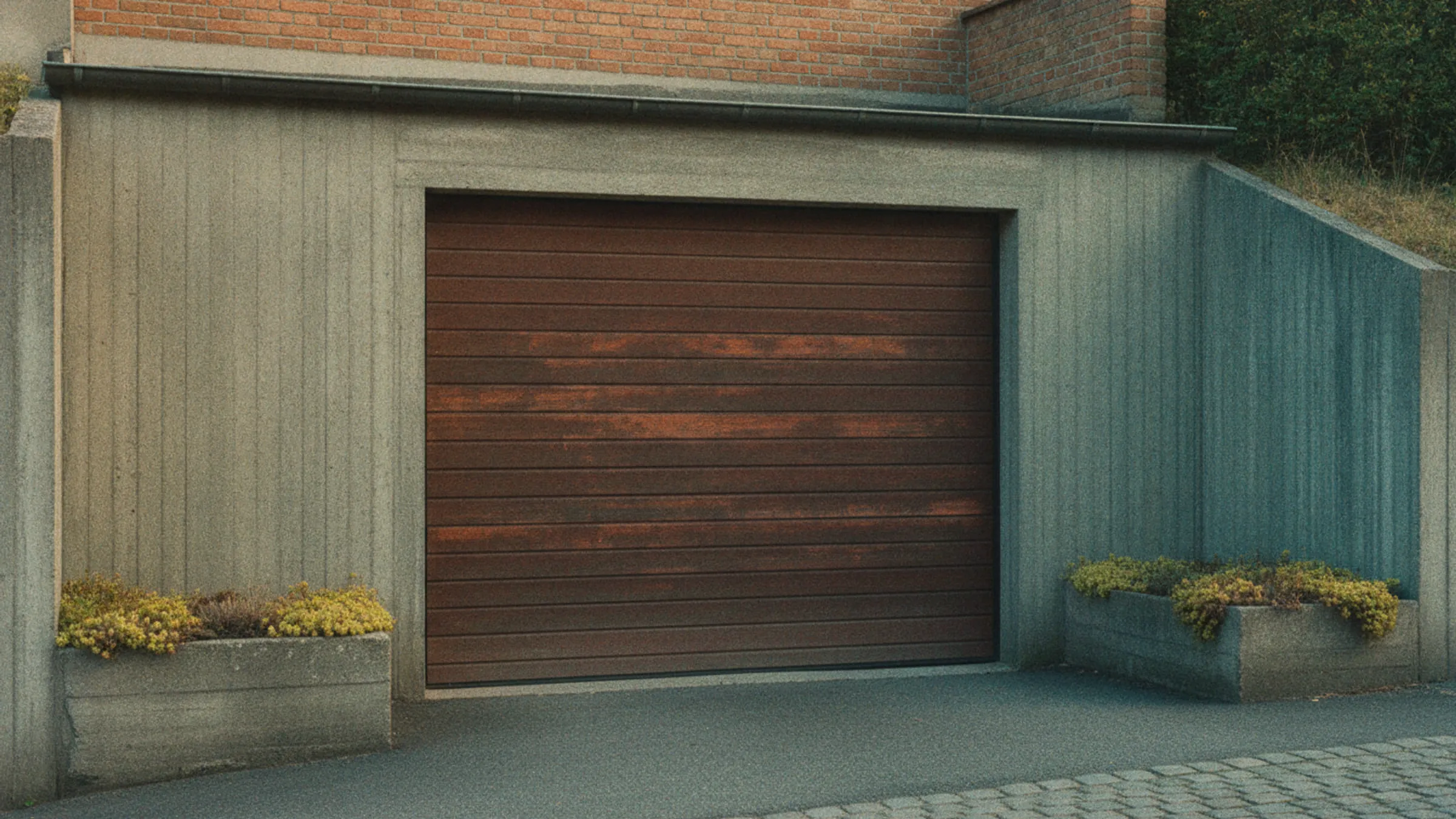 Garage indbygget i skråningen under et hus, med en betonstøttemur på begge sider og en rampe op til porten.