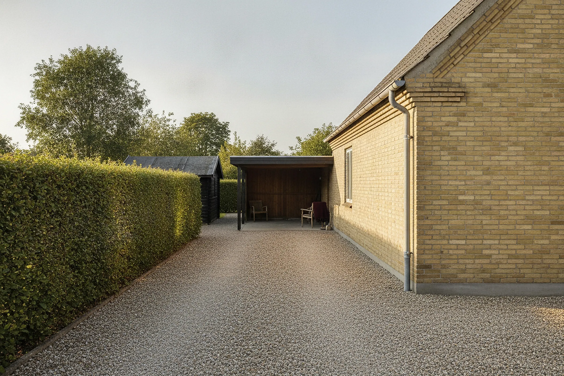 Grusindkørsel langs gavlen på et gult parcelhus med klippet ligusterhæk, carport og skur for enden i lav eftermiddagssol.