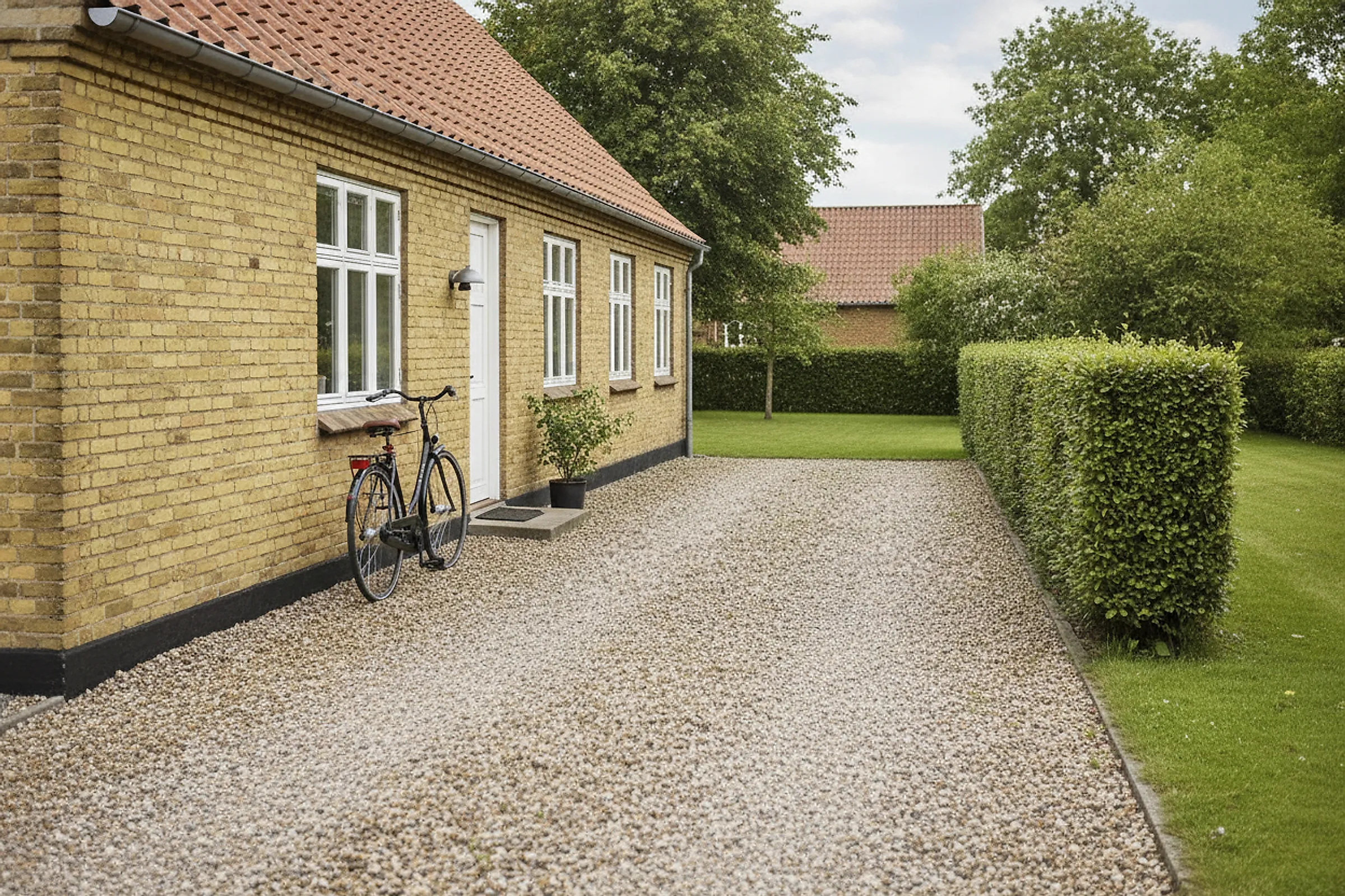 Gult parcelhus set skråt fra grusindkørslen i lav eftermiddagssol med klippet ligusterhæk og en cykel op ad muren.