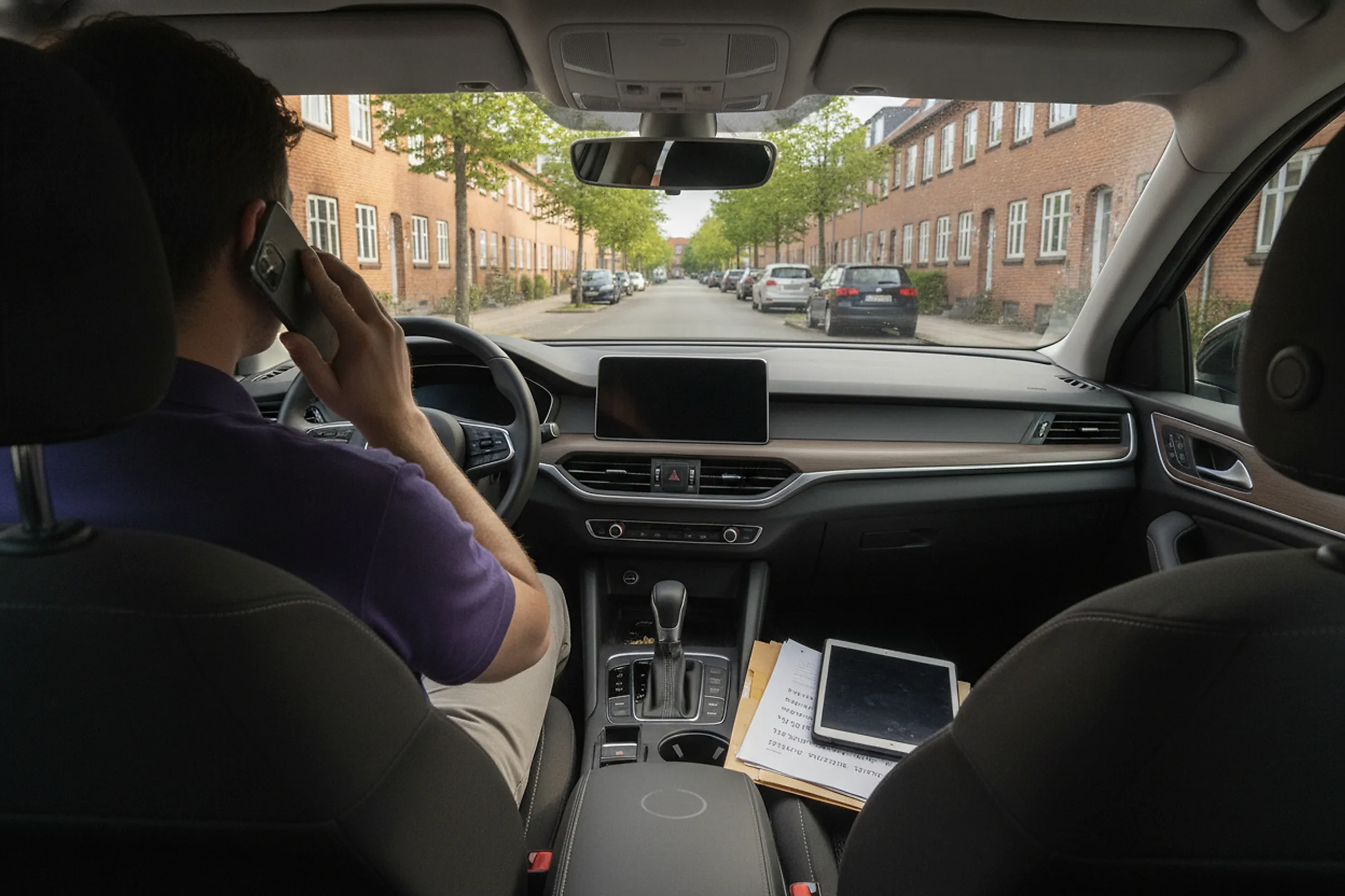 Partner sidder i en bil på en dansk villavej og telefonerer. På sædet ved siden af ligger papirer og en tablet.