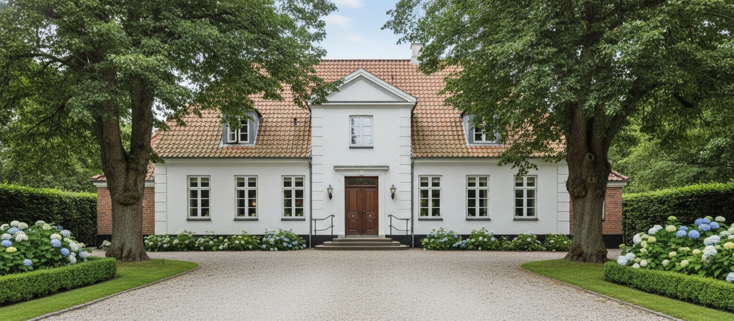 Hvid villa med rødt tegltag og frontispice for enden af en grusindkørsel mellem to store træer.