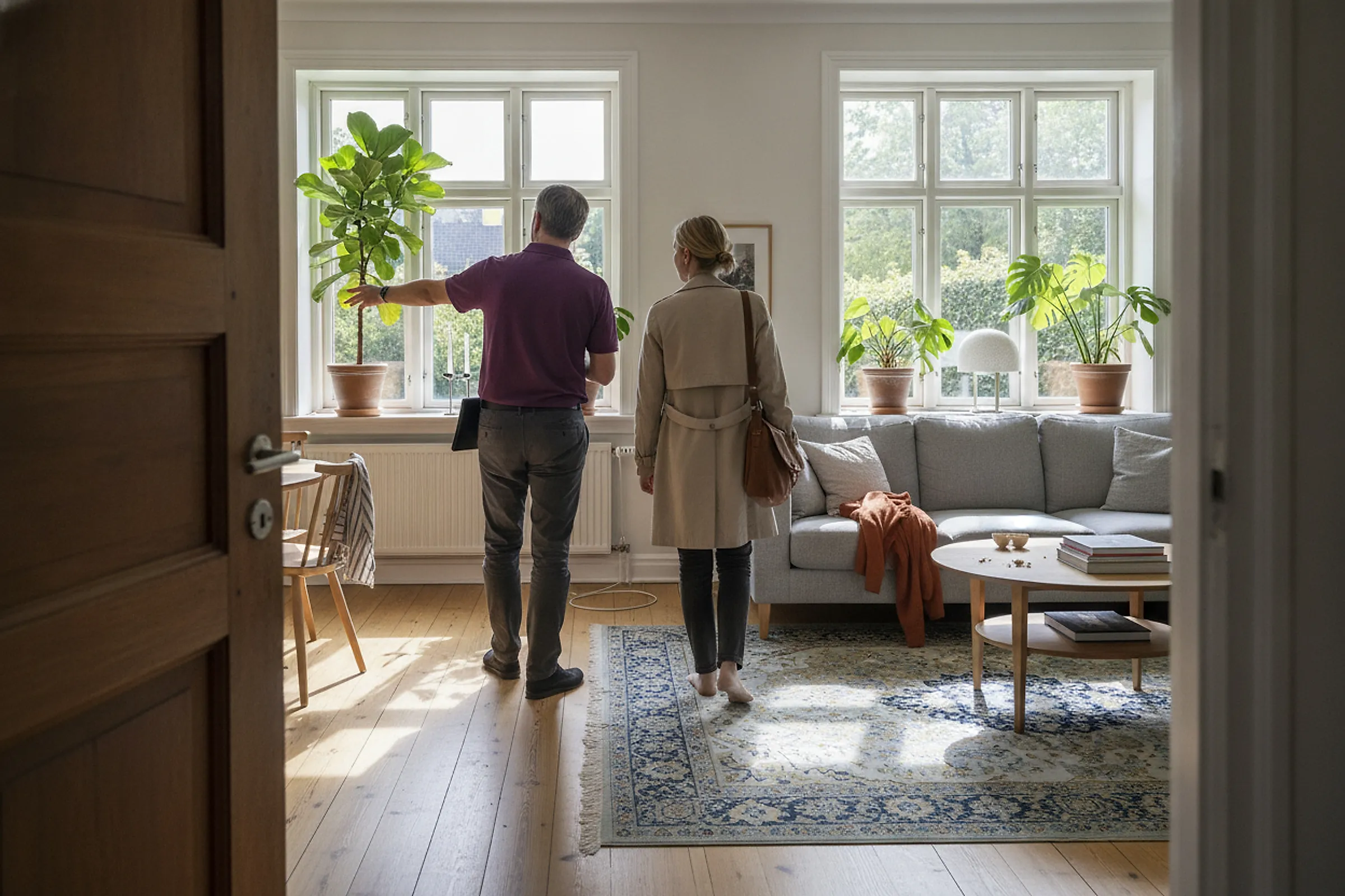 Partner viser en bolig frem for en interesseret køber i en lys stue med store vinduer og planter.